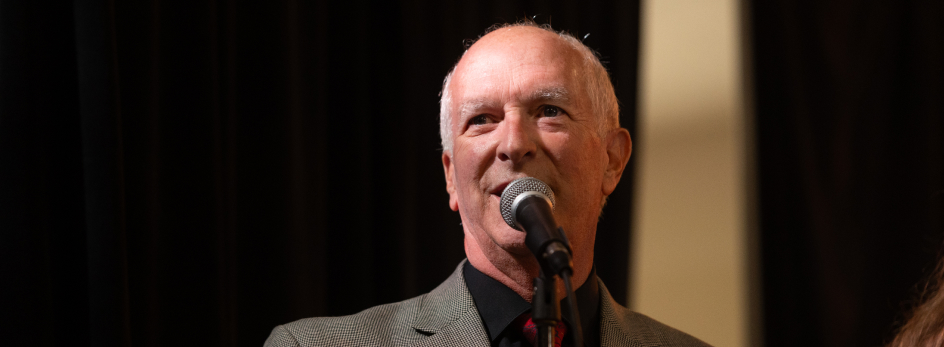 Dr. Rob Klevan speaking into a microphone in front of an out of focus black curtain