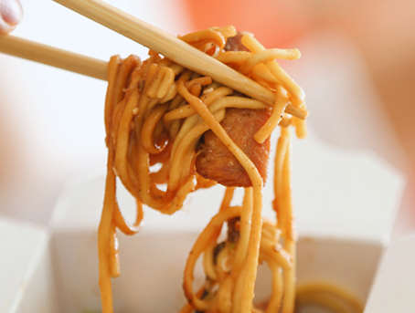 A pair of chopsticks taking noodles out of a take out box