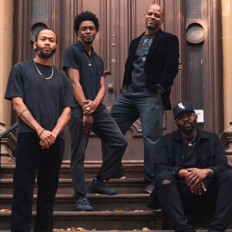 Three members of Upendo standing and one sitting on stairs in front of an outside door