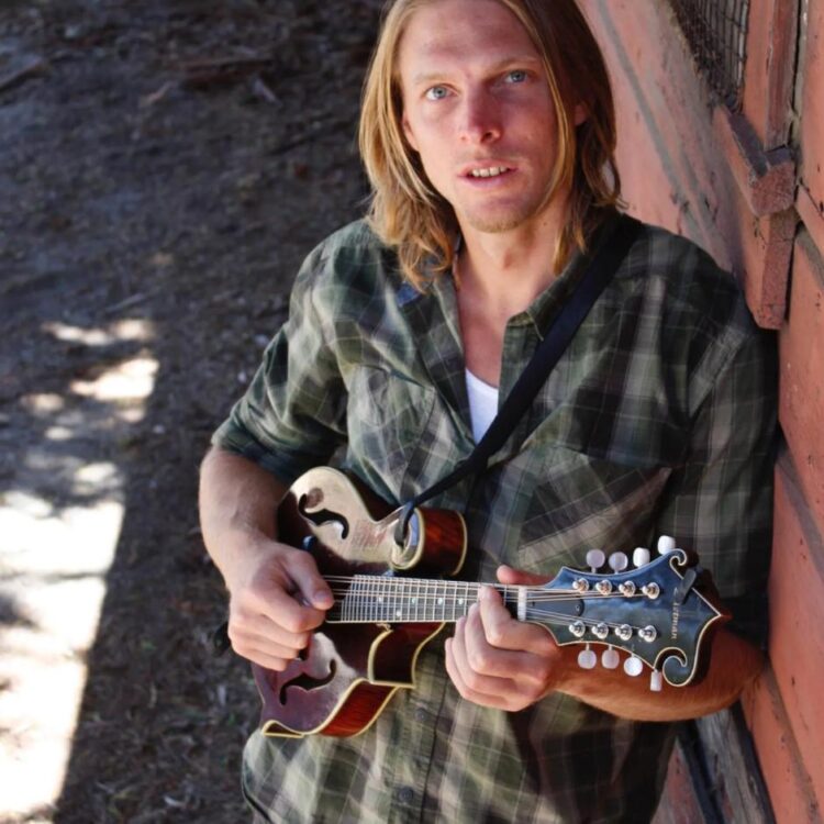 David Holodiloff holding a mandolin instrument leaning against a brick wall