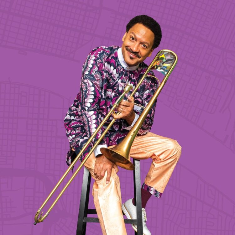 Delfeayo Marsalis sitting on a stool with his trombone in front of a light purple background