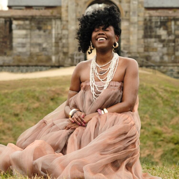 Ekep Nkwelle sitting on grass in front of an out of focus brick building