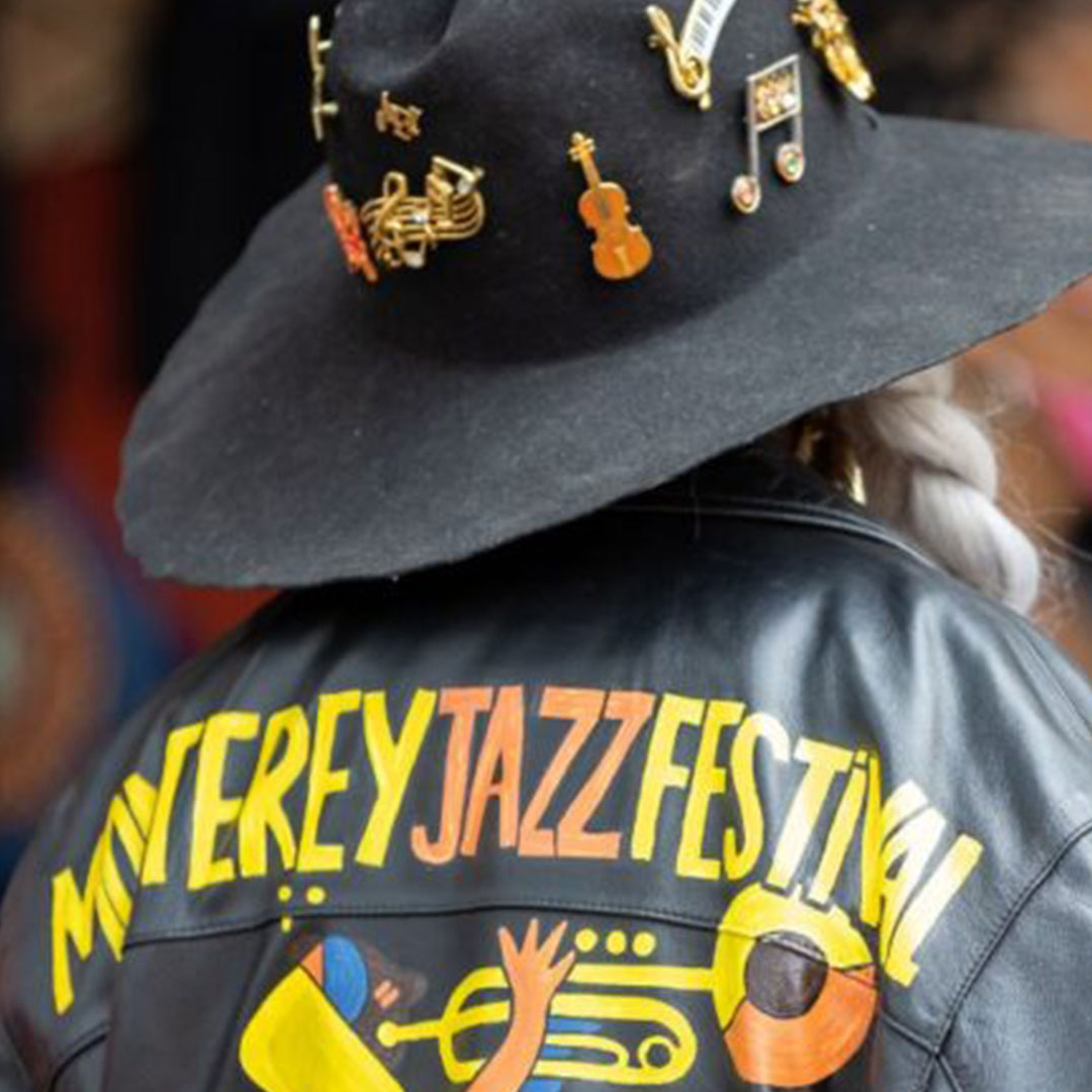 The back of someone wearing a black leather jacket that says 'Monterey jazz Festival' and a large brimmed hat with music note pins.