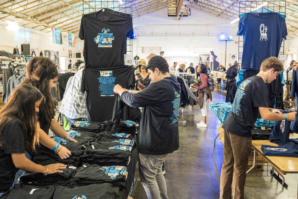 Merch room with volunteers organizing Monterey Jazz Festival merch on tables, with more racks and guests in the background