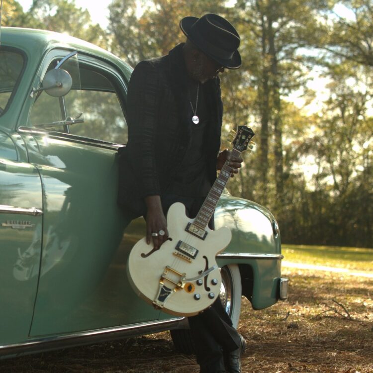 Mr. Sipp leaning against a retro light green car. Holding a white guitar against his legs.