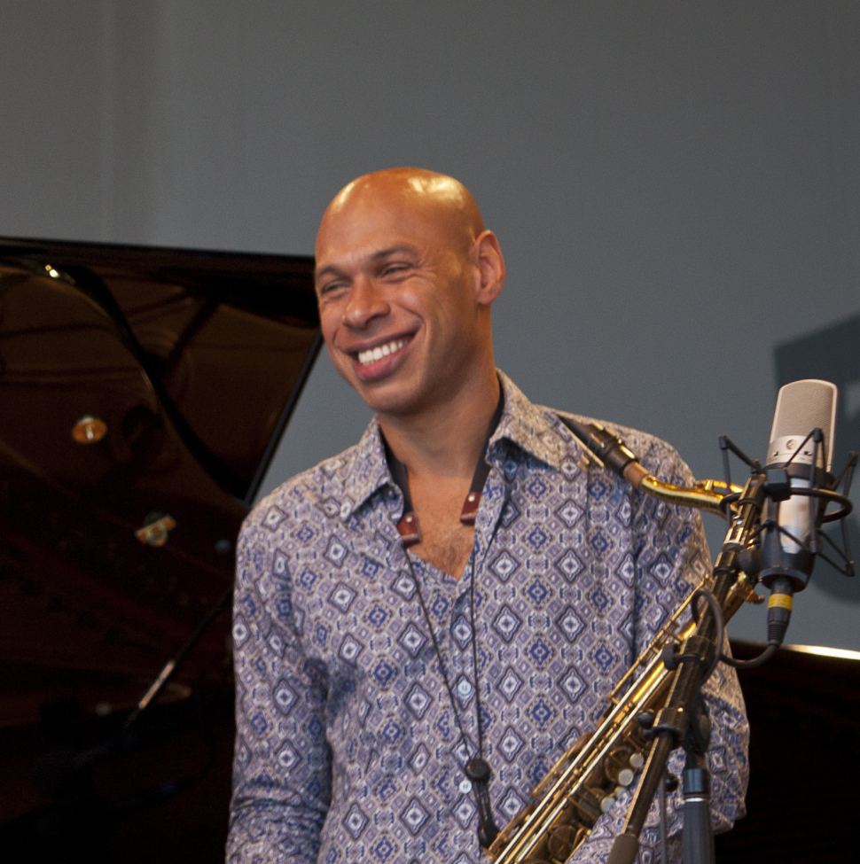 Joshua Redman holding a saxophone in front of an out of focus piano and light blue wall