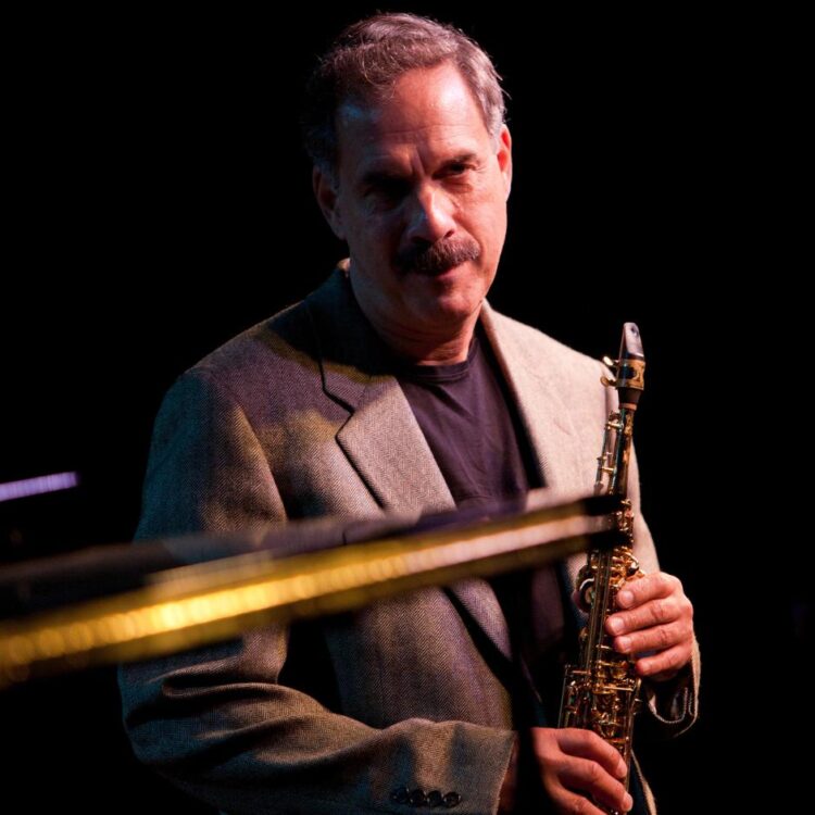 Paul Contos standing with his saxophone on stage, with an out of focus instrument in front of him
