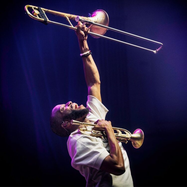 Trombone Shorty holding his trumpet to his chest with one arm, while yelling with his trombone held in the air with the other arm