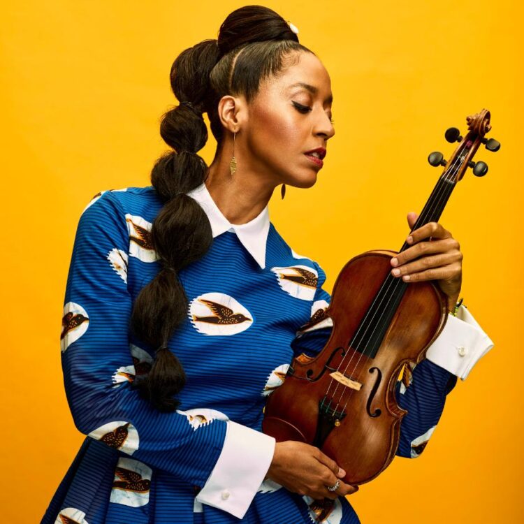 Yilian Cañizares standing with her violin in front of an orange-yellow background