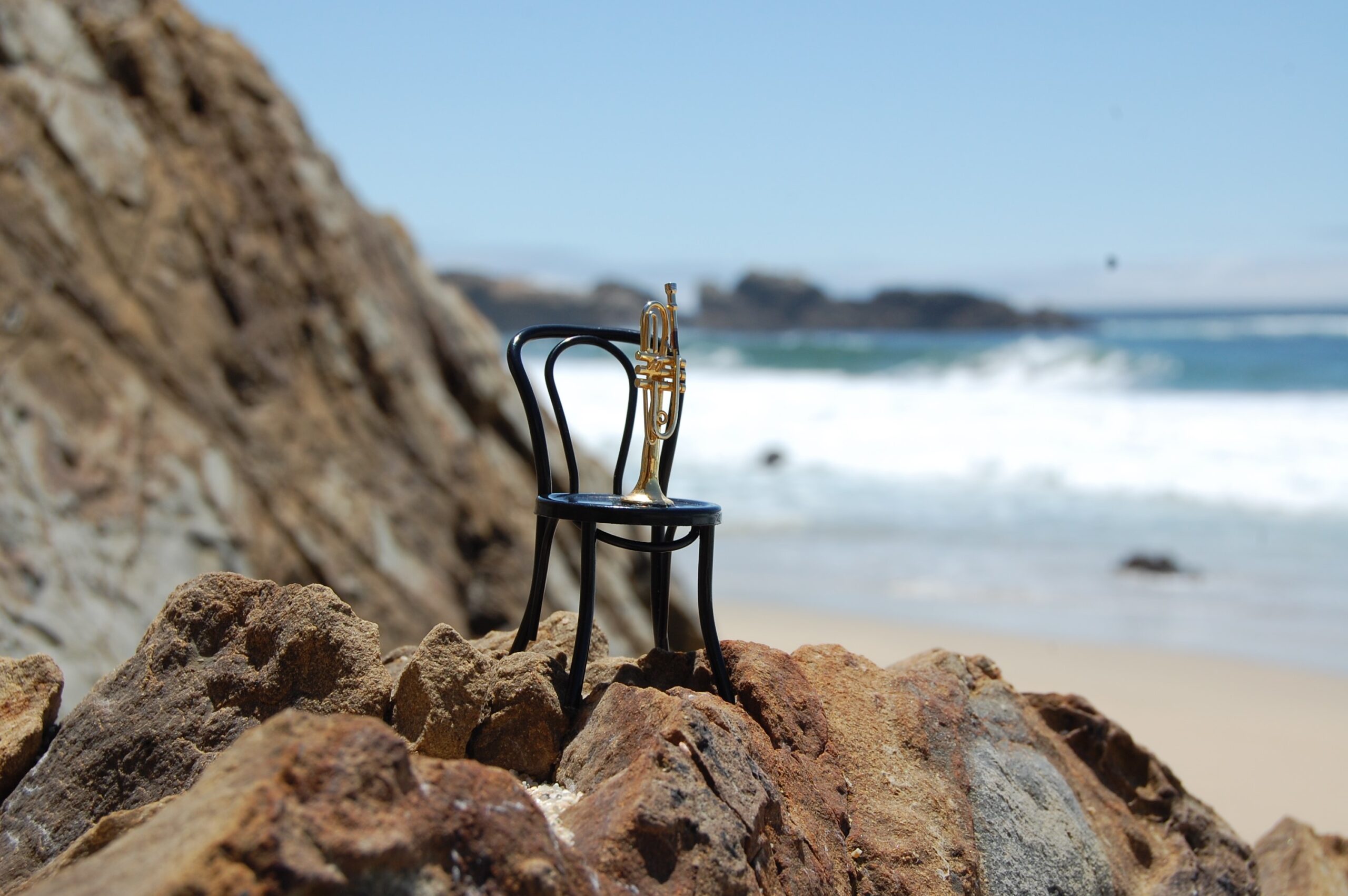 Black chair with a trumpet on the seat. Placed on some rocks with a beach in the background.