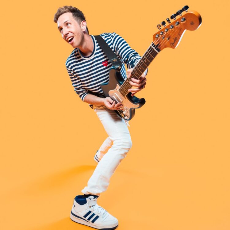 Cory Wong standing with his guitar in front of an orange yellow background