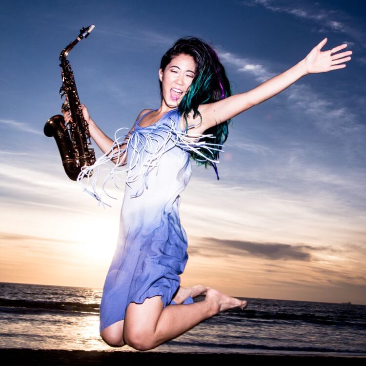 Grace Kelly holding her saxophone, jumping in the air with arms out stretched in front of the ocean